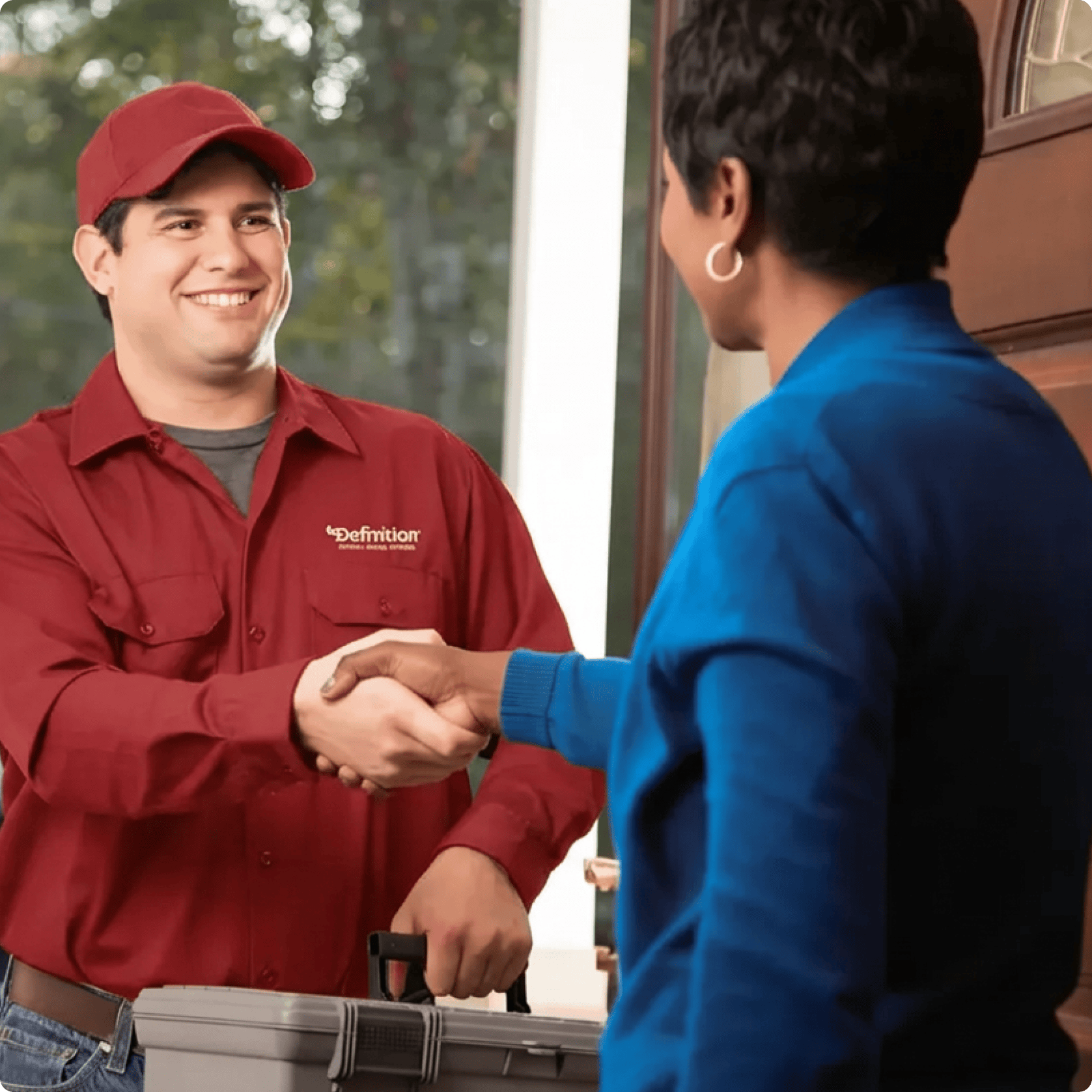 A Definition Heating, Cooling & Plumbing technician in a maroon uniform and cap shaking hands with a female homeowner at the front door, providing friendly in-home service and customer support.